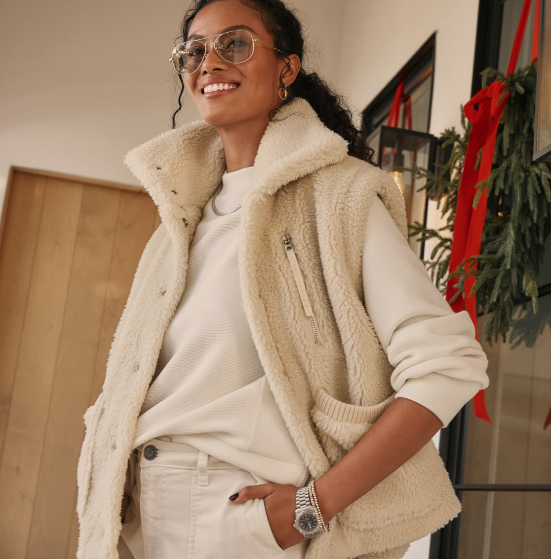 Woman stands near a holiday wreath wearing an ivory sweatshirt, an ivory faux-sherpa vest, and a pair of ivory pants.