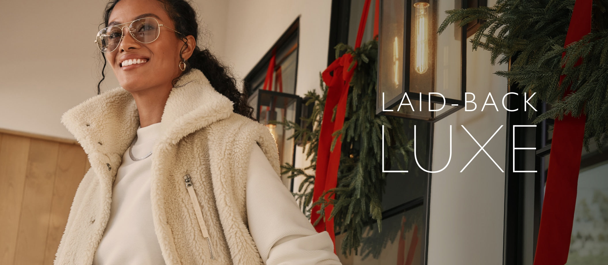 Woman standing outside near a holiday wreath wears an ivory sherpa vest over an ivory sweatshirt. Text over image reads: Laid-Back Luxe